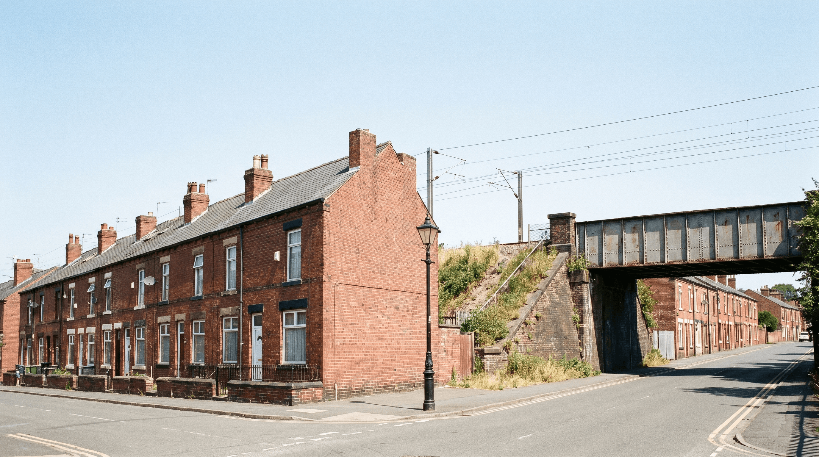 Row of Edwardian red-brick terraced houses ending at a railway embankment with overhead electrification wires and a steel railway bridge in the mid-distance, in a UK city on a bright summer day